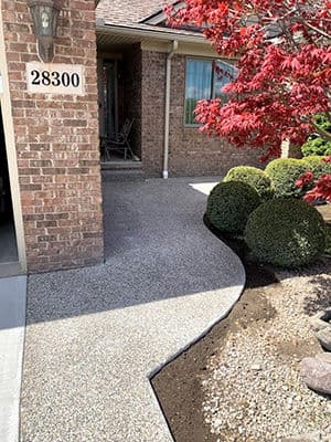 Curved gravel walkway leading to a brick house with manicured bushes and a red maple tree.