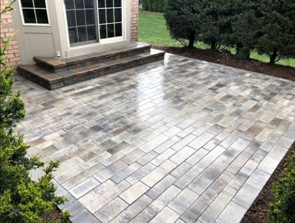 Patio with gray and beige stone pavers, featuring steps and green landscaping nearby.