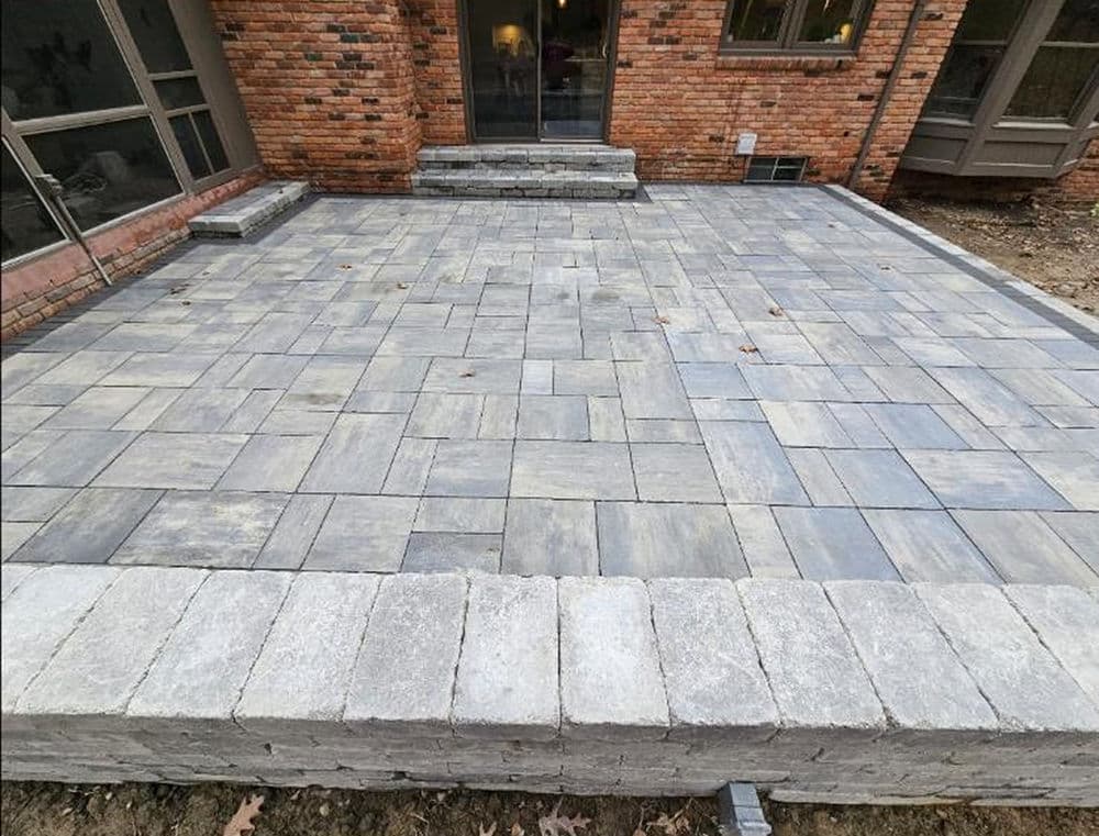 Paver patio installation with gray stones, adjacent to a brick house and steps.