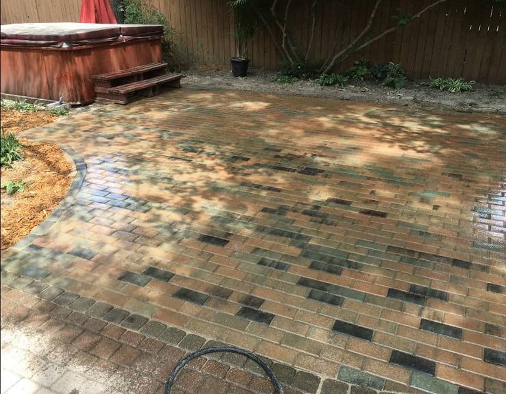 Paved backyard patio with a hot tub and lush greenery in the background.