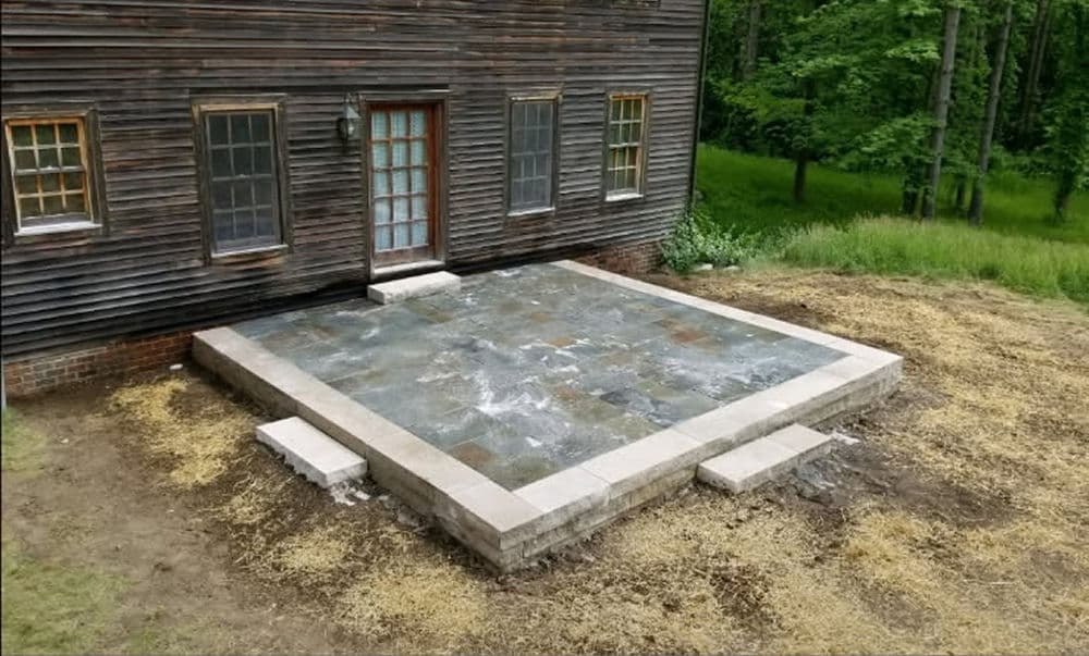 Newly installed stone patio next to a rustic wooden house, surrounded by greenery.