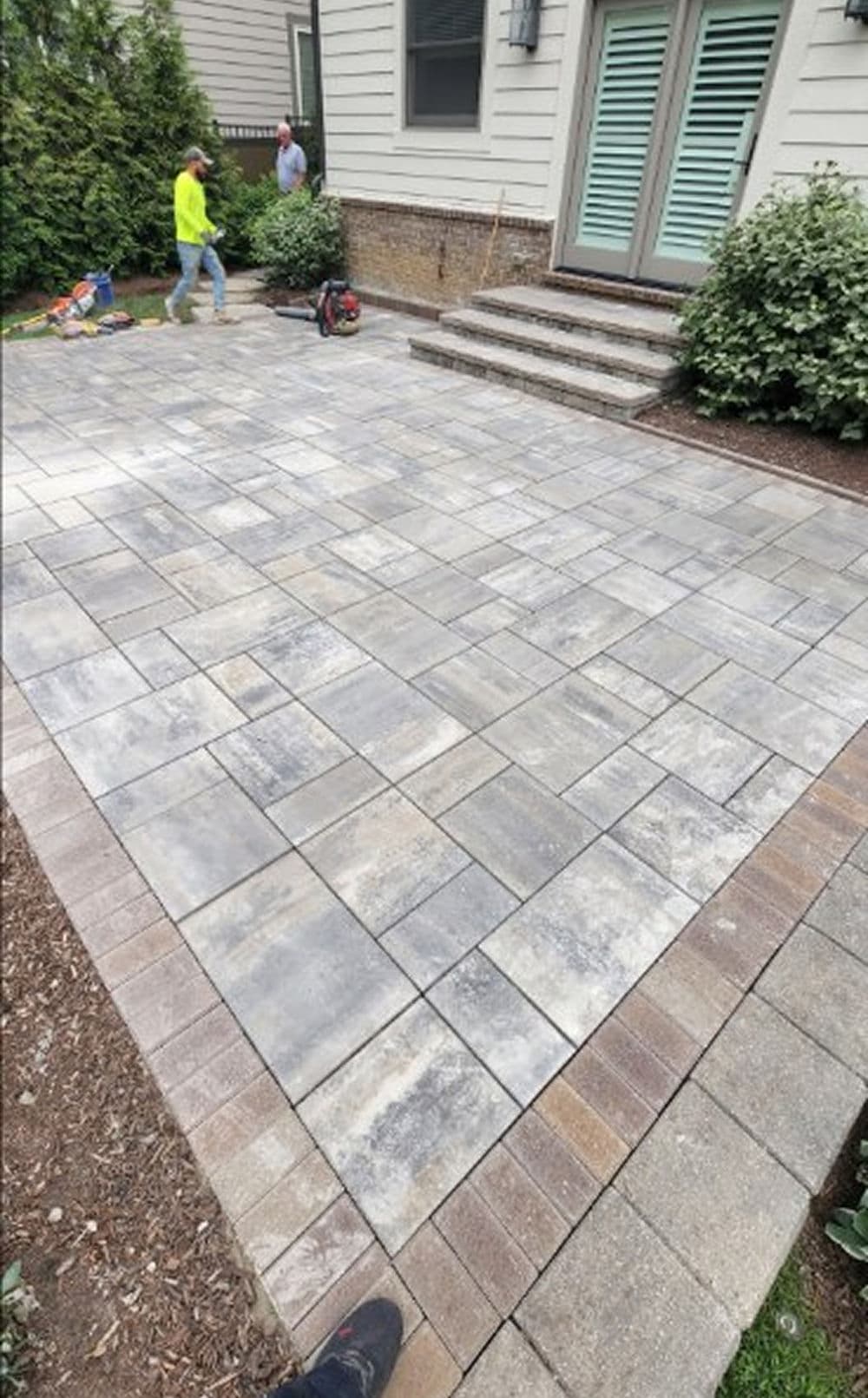 Newly installed stone patio with a worker in the background and landscaped greenery.