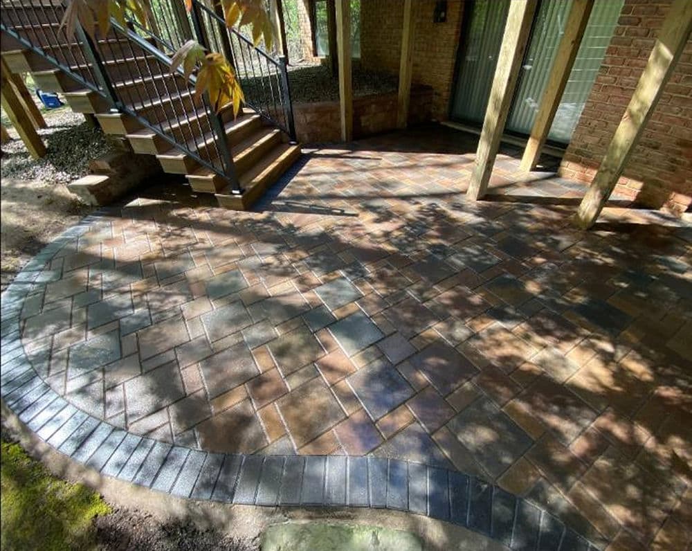 Paved outdoor patio with a herringbone pattern and wooden stairs in a wooded setting.