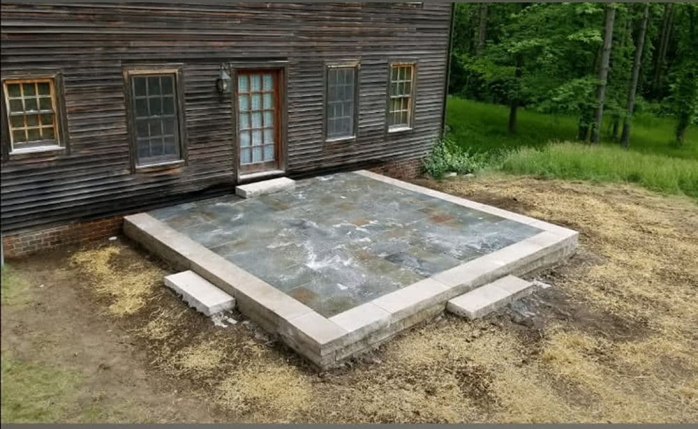 Newly constructed stone patio in front of rustic wooden house, surrounded by greenery.