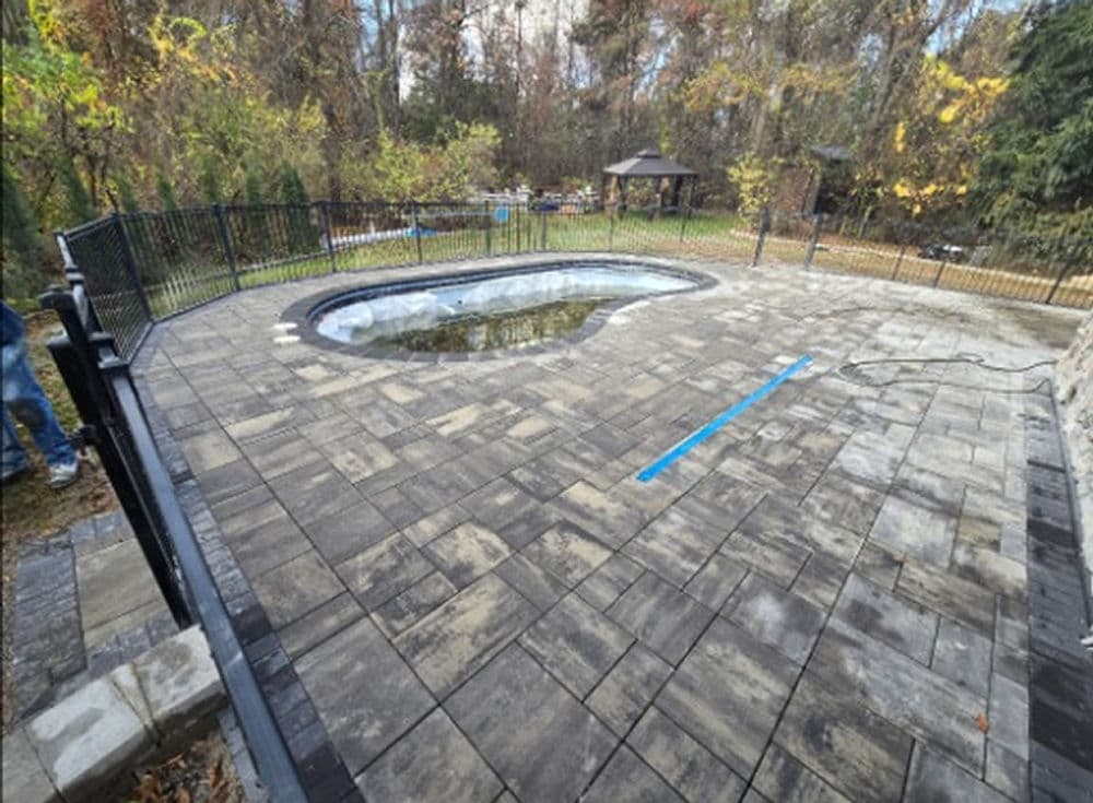 In-ground pool surrounded by gray paver patio and black fence in a wooded backyard setting.