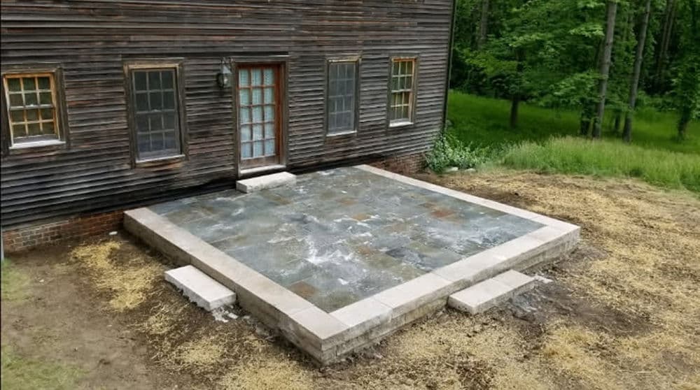 Slate patio under construction next to a rustic wooden house surrounded by greenery.