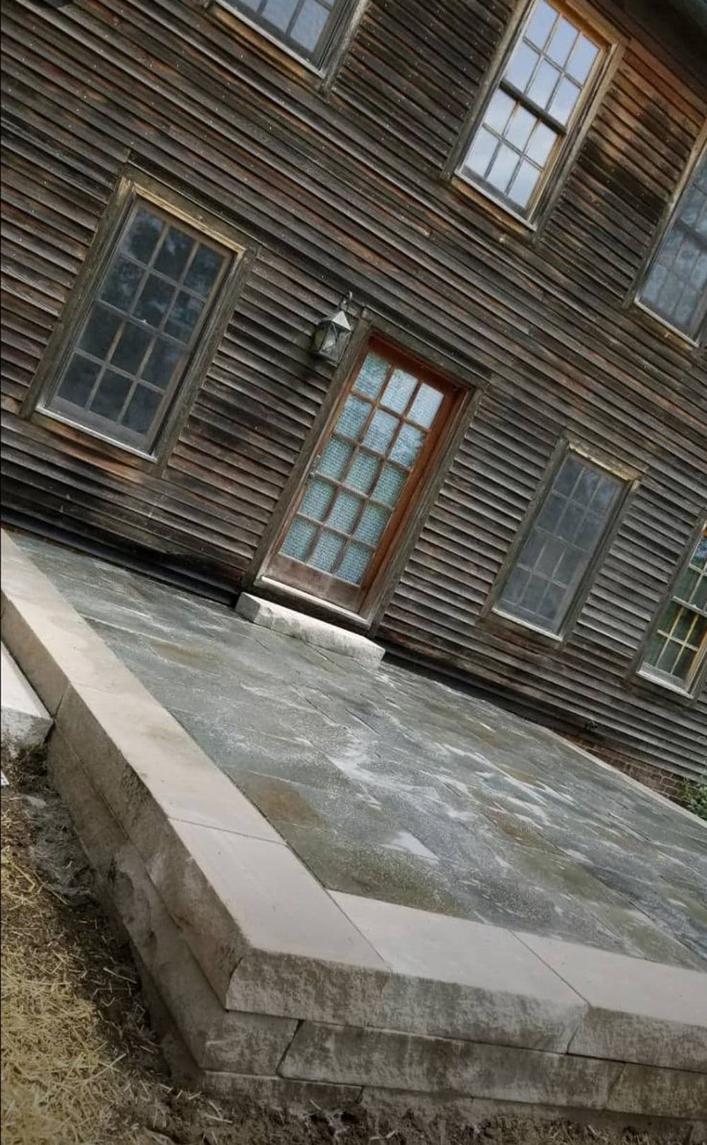 Historic wooden house with stone patio and large glass door, surrounded by natural landscaping.