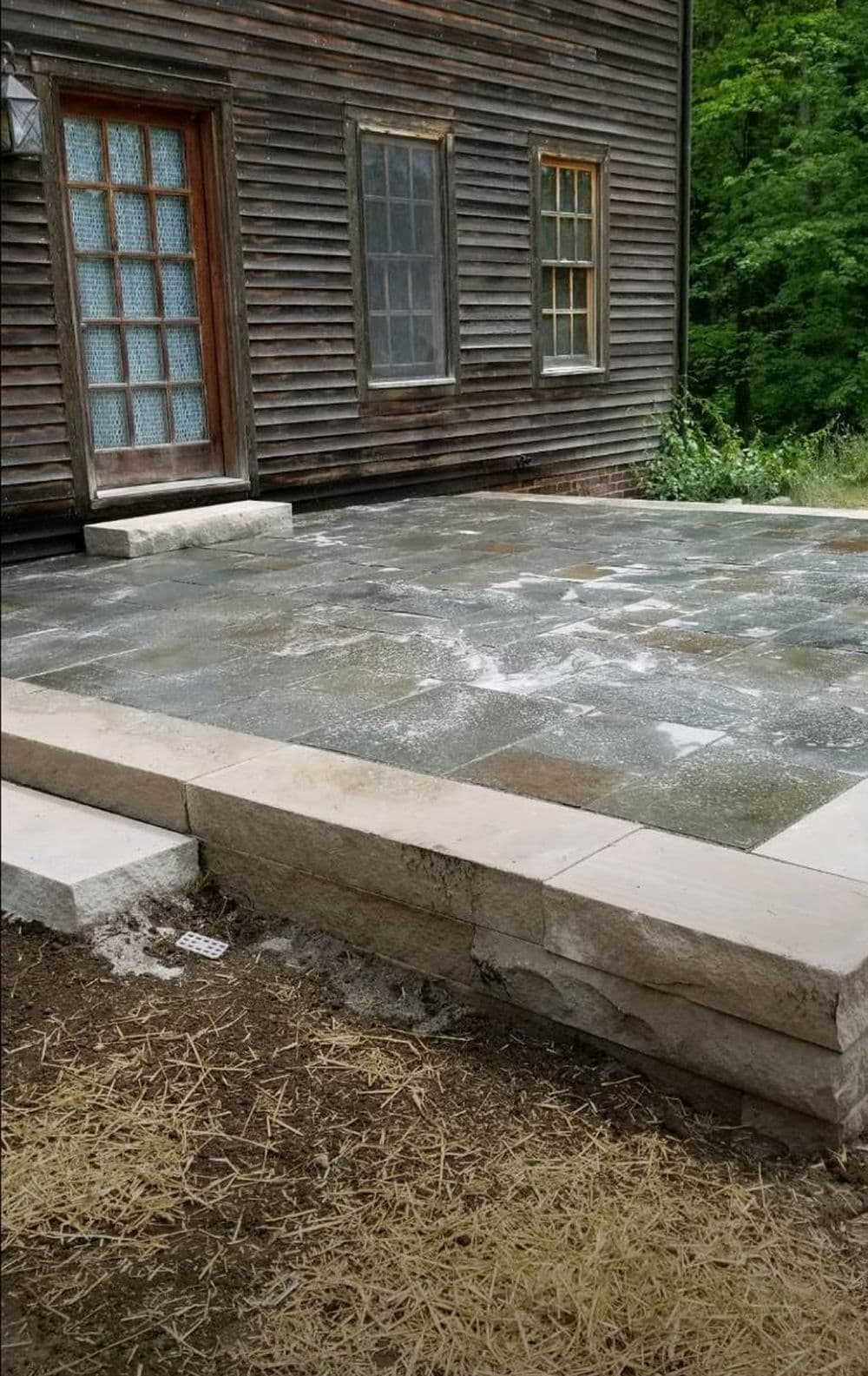 Newly constructed stone patio beside a rustic wooden house, surrounded by greenery.